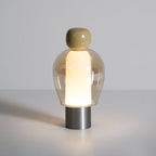 Lit Clérachic table lamp on a pale surface, an olive cap and amber glass dome above a brushed steel base, with a soft glow below.
