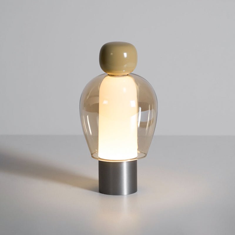 Lit Clérachic table lamp on a pale surface, an olive cap and amber glass dome above a brushed steel base, with a soft glow below.