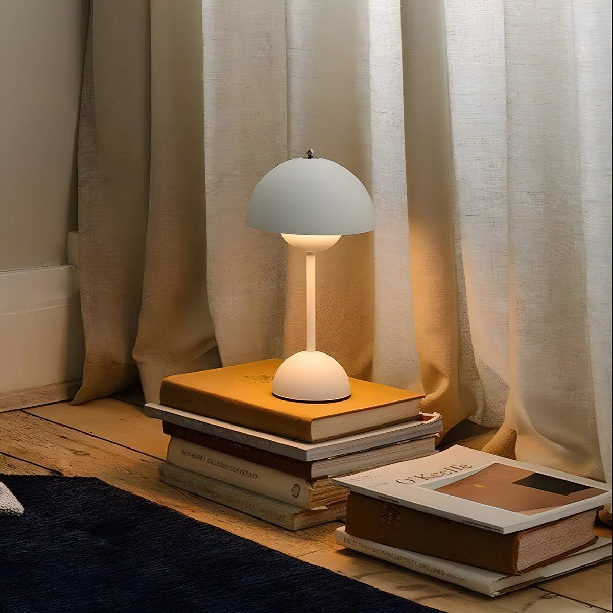 A pale grey Clérachic table lamp glowing softly on a stack of books beside a sheer linen curtain.
