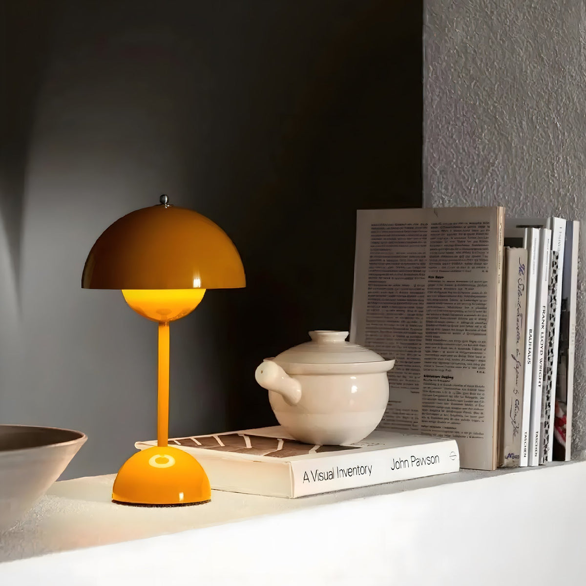 A mustard-yellow Clérachic table lamp lit warm on a white shelf, next to a ceramic pot and a row of books.
