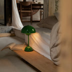 A green Clérachic table lamp lit beside a bed, with an open book resting on the low wooden bedside shelf.