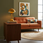 A small mustard Clérachic table lamp on a walnut sideboard, in a living room with an orange sofa and olive walls.