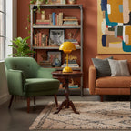 A yellow Clérachic table lamp on a round side table, set between a green armchair and a tall bookshelf.