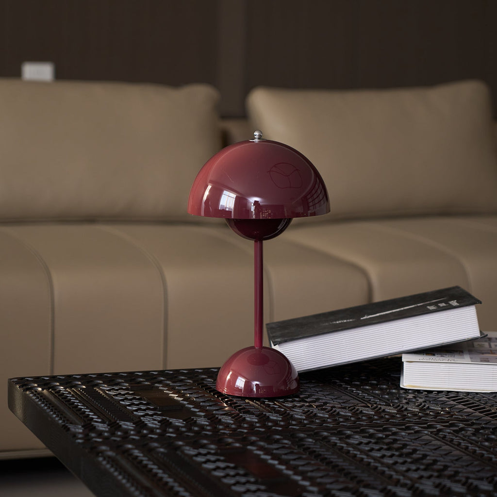 A burgundy Clérachic table lamp on a dark patterned table in front of a beige sofa, with a book beside it.