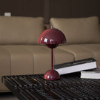 A burgundy Clérachic table lamp on a dark patterned table in front of a beige sofa, with a book beside it.