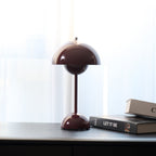 This Clérachic table lamp in deep plum on a dark desk, with two books and a bright curtain behind.
