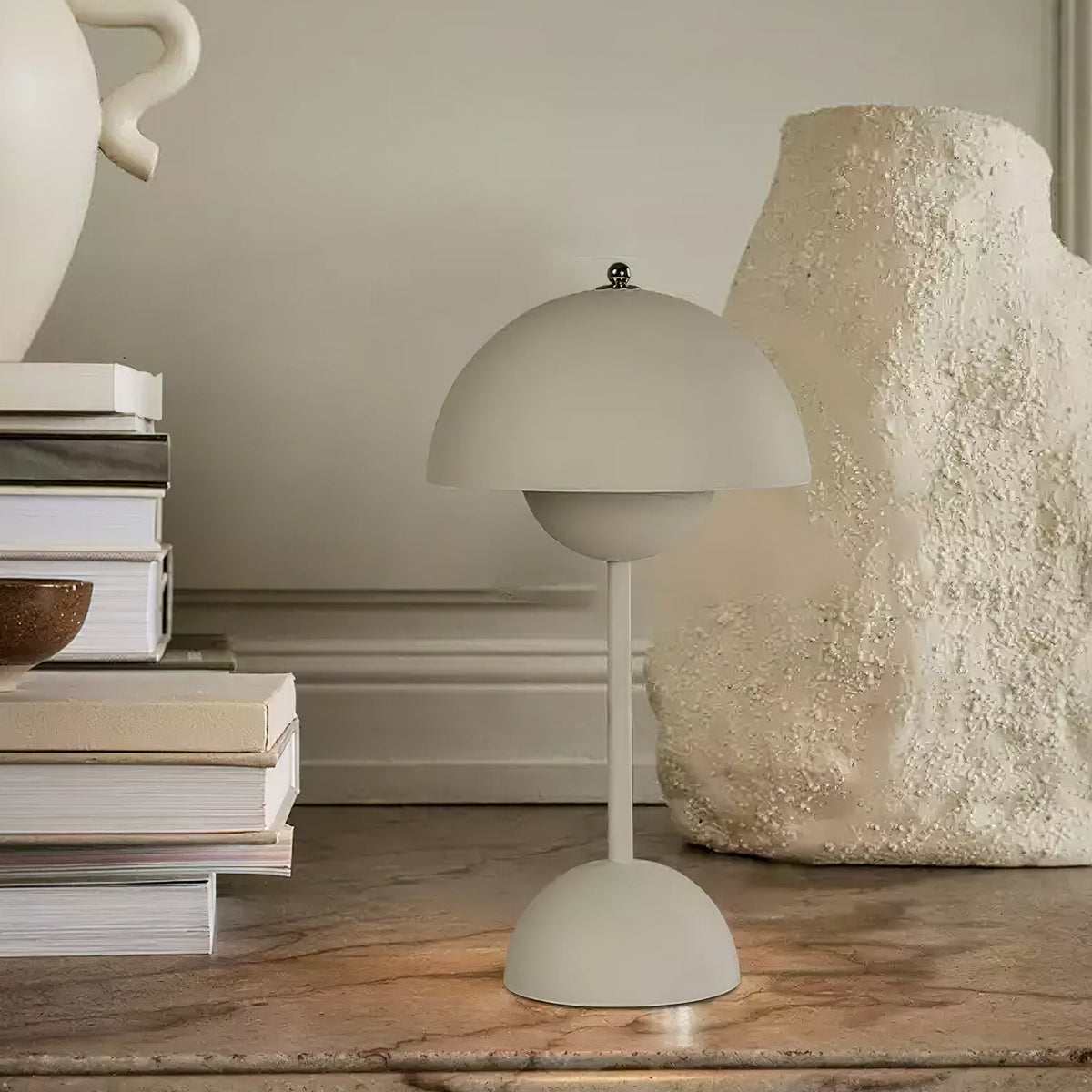 A matt grey-beige Clérachic table lamp on a marble shelf, beside a stack of books and a pale sculptural vase.