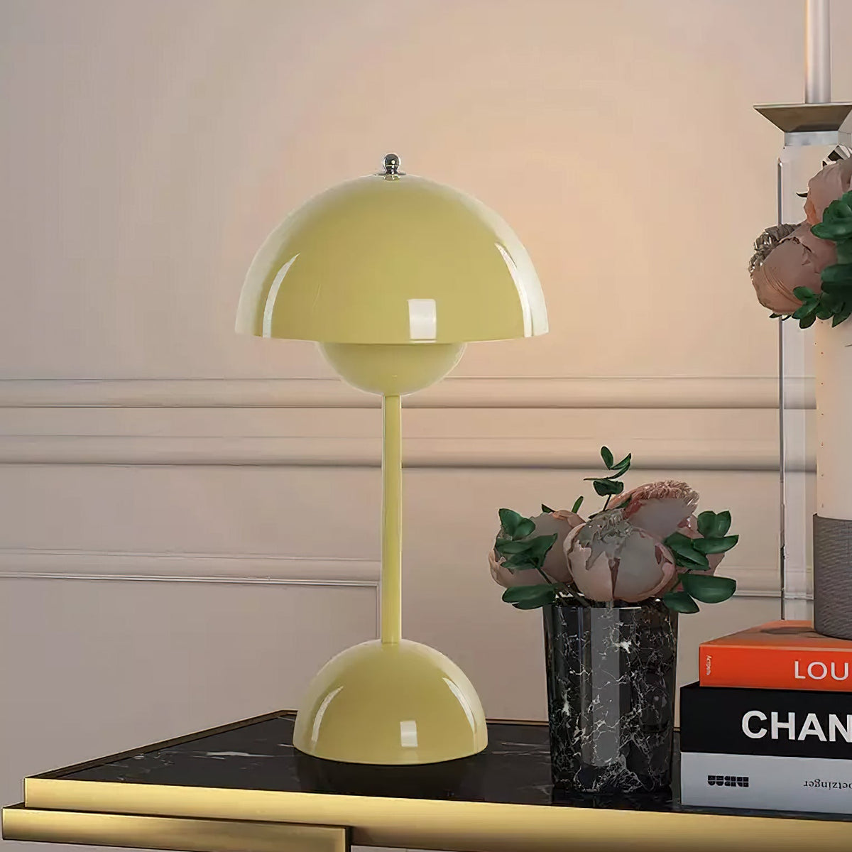 A pale yellow Clérachic table lamp on a gold-edged side table, next to a small vase of pink flowers.