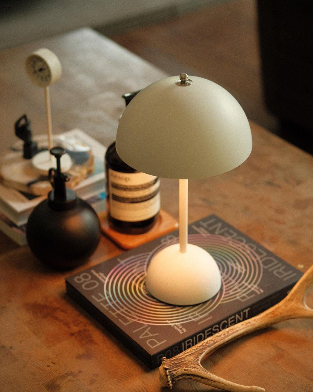 Close view of a cream Clérachic table lamp on a stack of books, with a small clock and a black bottle behind it.