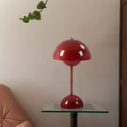 A red Clérachic table lamp on a round glass side table against a warm beige wall, with a trailing leaf above.