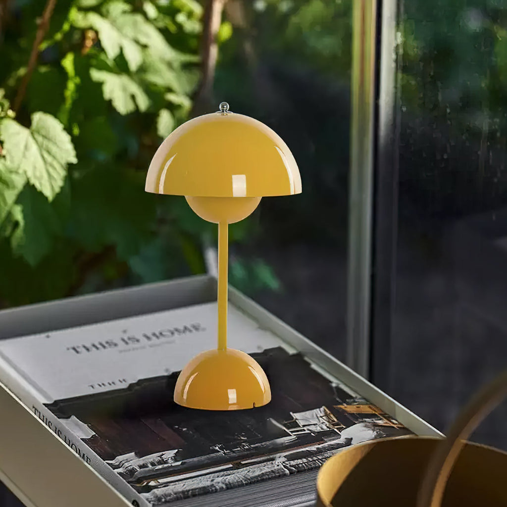 This Clérachic table lamp in yellow on a metal tray with a coffee-table book, beside a window with garden leaves.