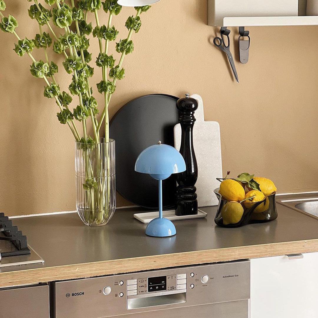 A light-blue Clérachic table lamp on a kitchen worktop, beside a glass vase, a pepper mill and a bowl of lemons.