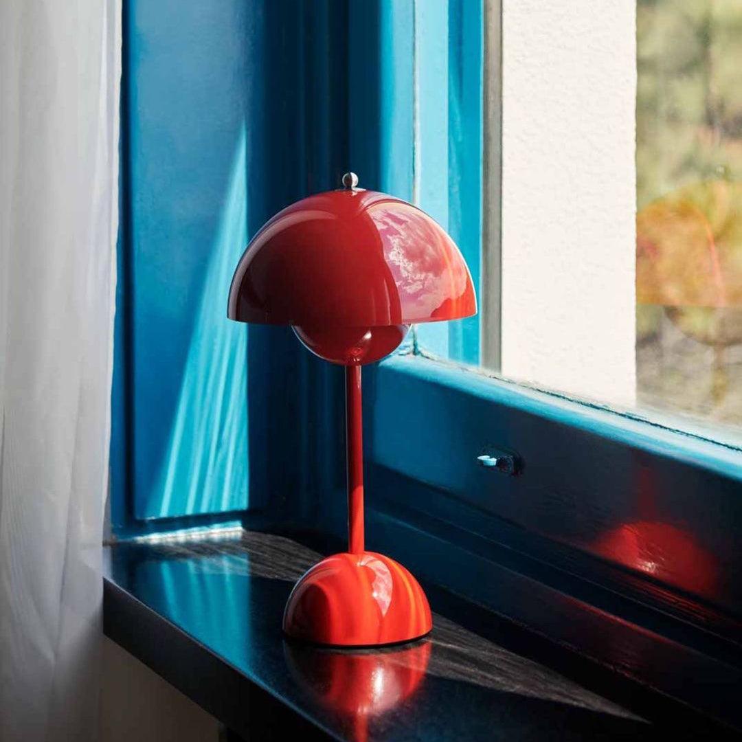 A red Clérachic table lamp on a dark windowsill, with a bright blue window frame and a white curtain behind.