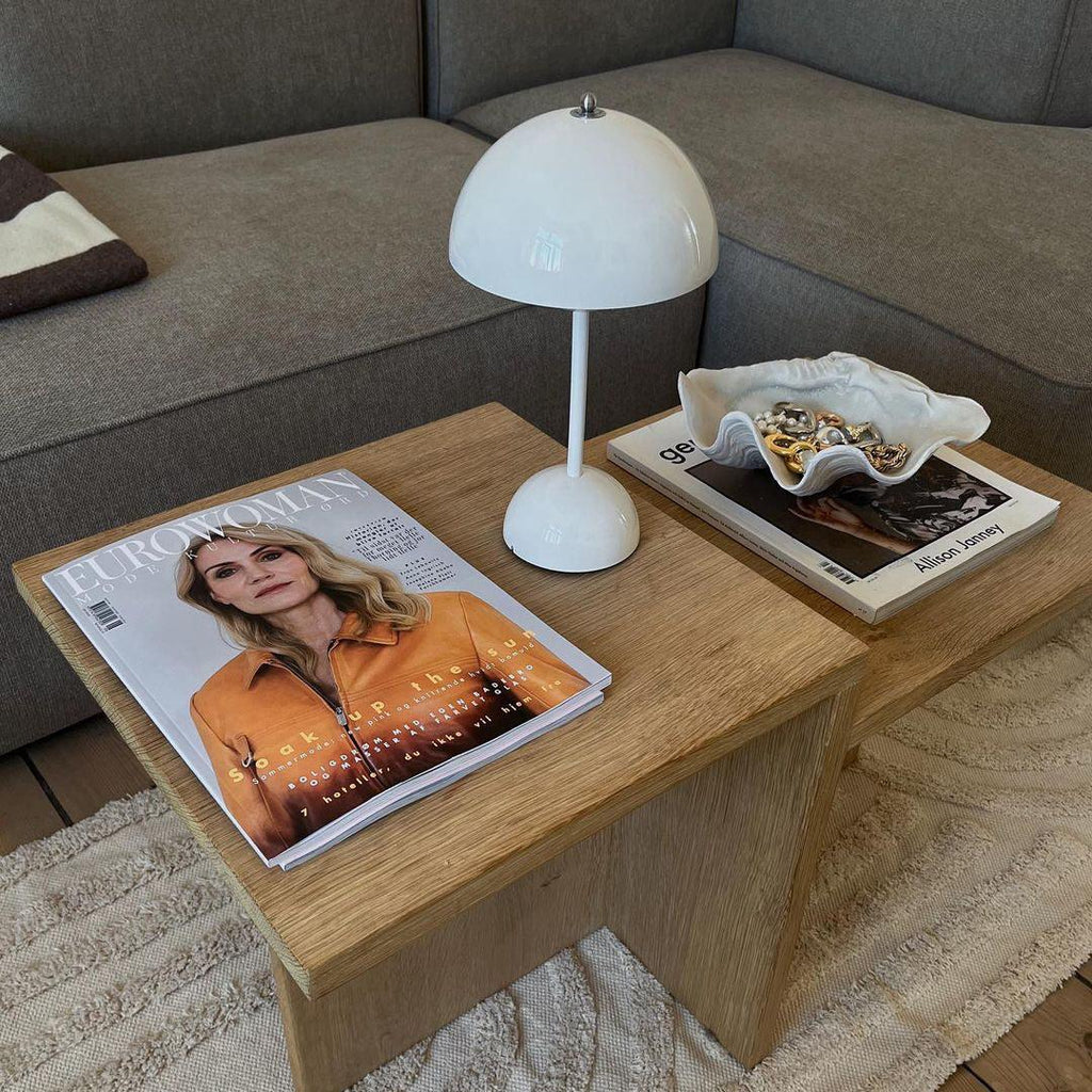 A white Clérachic table lamp on a wooden coffee table with magazines and a shell dish, in front of a grey sofa.