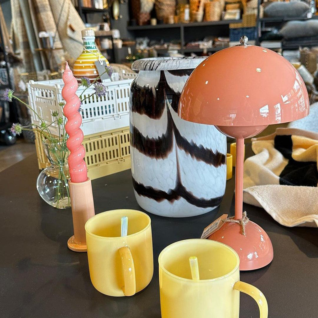 A coral-pink Clérachic table lamp on a shop counter among glassware, yellow mugs and a twisted pink candle.