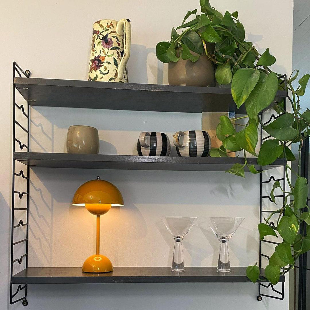 A yellow Clérachic table lamp lit on the bottom shelf of a dark wall unit, with cocktail glasses and a trailing plant.