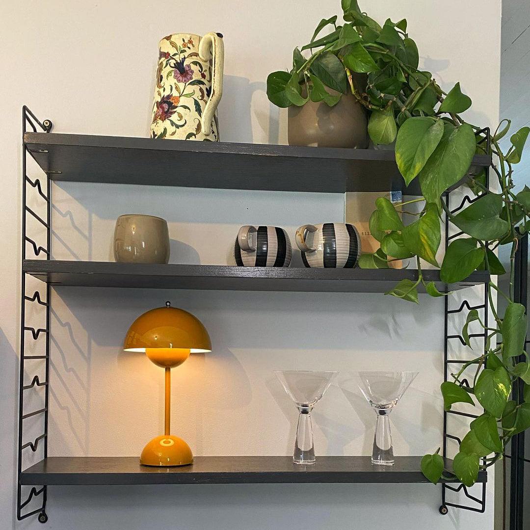 A yellow Clérachic table lamp lit on the bottom shelf of a dark wall unit, with cocktail glasses and a trailing plant.