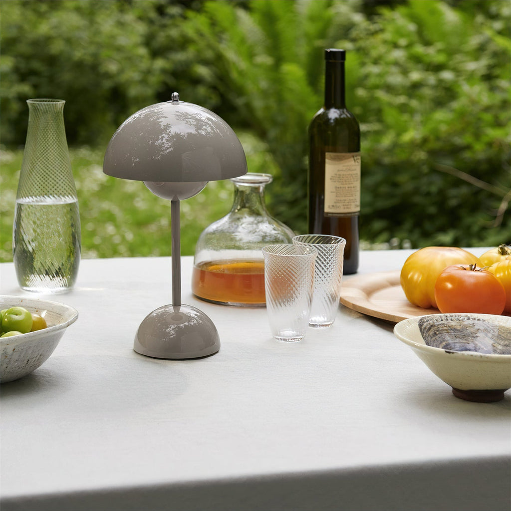 A grey Clérachic table lamp on an outdoor table with carafes, glasses and tomatoes, set against green foliage.
