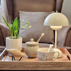 A cream Clérachic table lamp lit on a wooden tray with a plant pot, a honey jar and a cup of coffee.