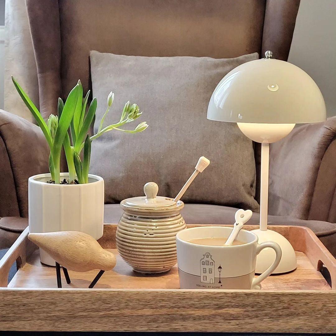 A cream Clérachic table lamp lit on a wooden tray with a plant pot, a honey jar and a cup of coffee.