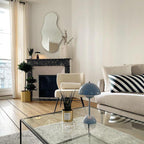 A pale blue Clérachic table lamp on a glass coffee table in a bright living room with a cream sofa and parquet floor.