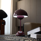 A deep plum Clérachic table lamp lit from within the shade, on a dark patterned table beside a hardback book.