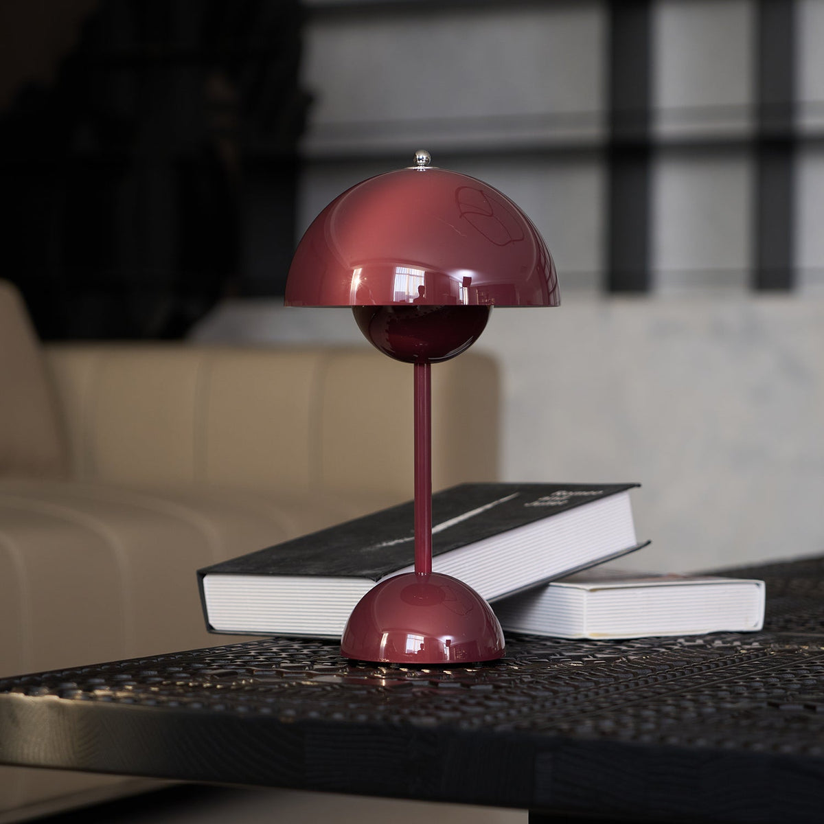 This Clérachic table lamp in burgundy with a dark book leaning against its base, in front of a beige sofa.
