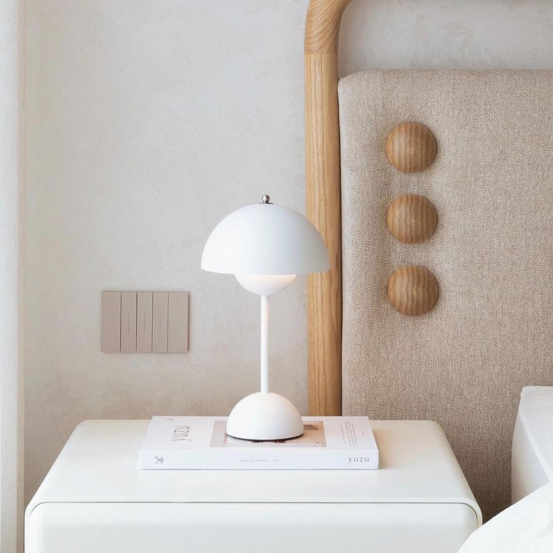 A white Clérachic table lamp on a white bedside table with a book, beside a rattan headboard with wooden pegs.