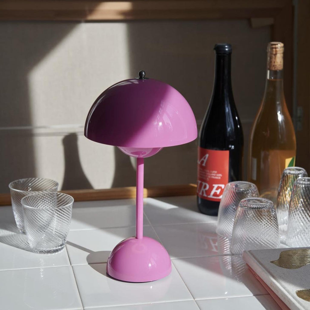 A magenta Clérachic table lamp on a white tiled surface in sunlight, with two bottles and glasses behind.