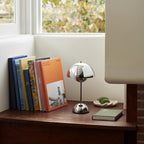 A small chrome-finish Clérachic table lamp on a wooden shelf, beside a row of books and a green dish.