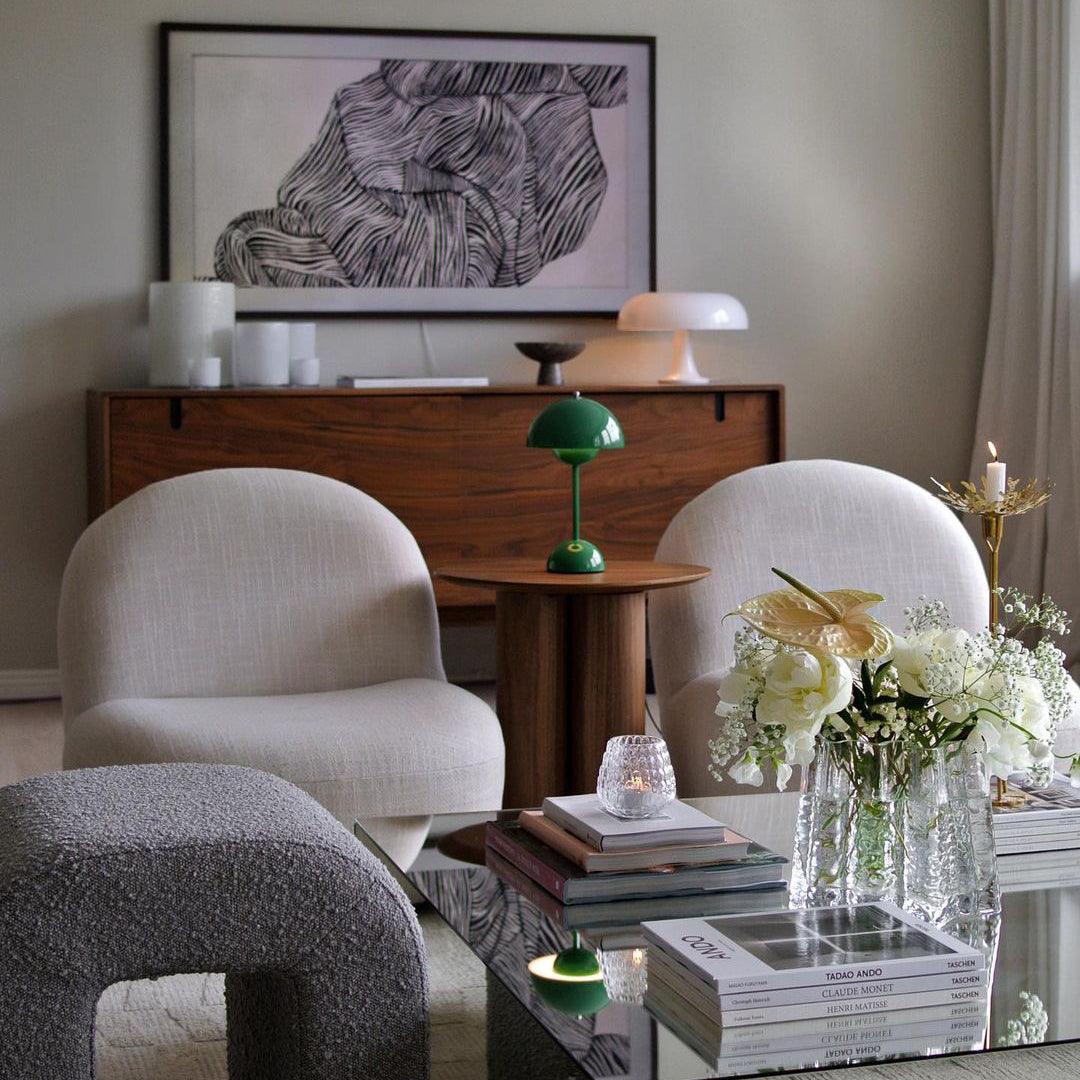 A small green Clérachic table lamp on a round wooden table in a living room with cream armchairs and a framed print.