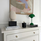 A green Clérachic table lamp on a white chest of drawers, beside design books, a candle and a plaster face.