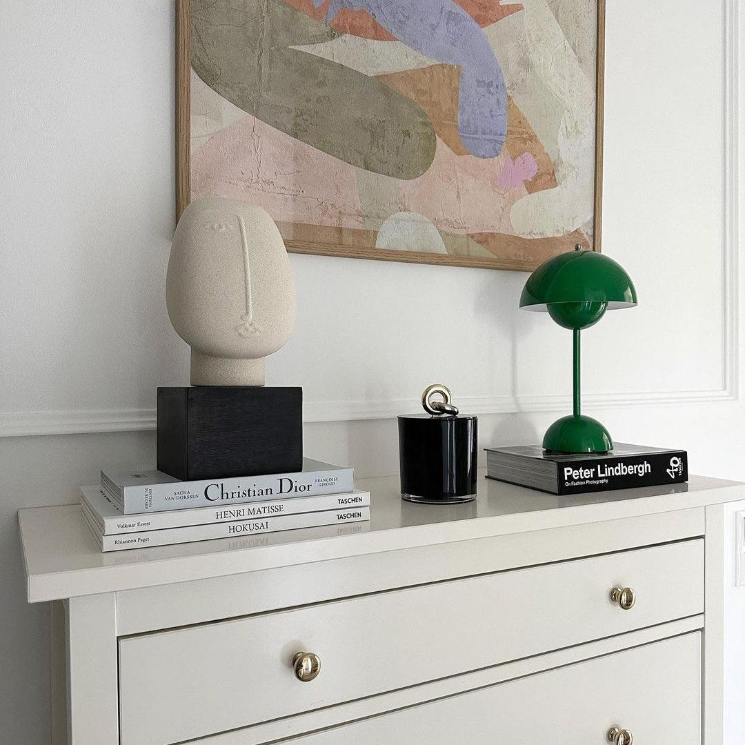 A green Clérachic table lamp on a white chest of drawers, beside design books, a candle and a plaster face.