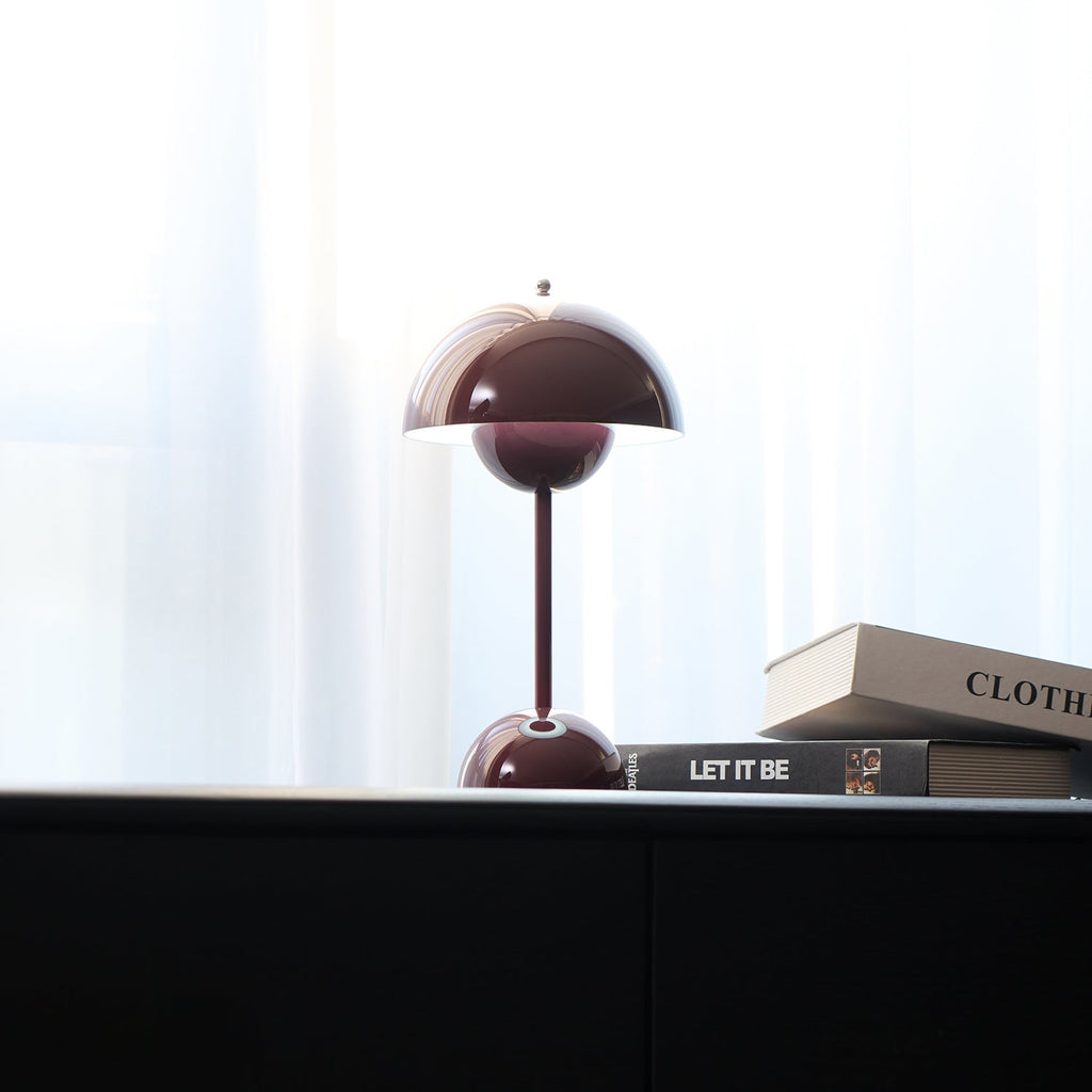 A deep plum Clérachic table lamp half-hidden behind books on a black cabinet, in front of a sheer white curtain.