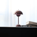 A deep plum Clérachic table lamp half-hidden behind books on a black cabinet, in front of a sheer white curtain.