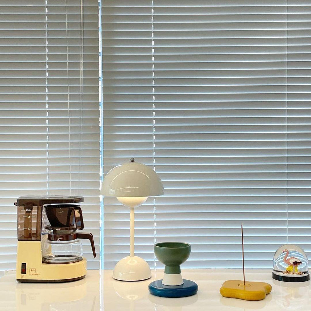 A pale cream Clérachic table lamp lit on a white shelf, beside a coffee maker and small coloured ornaments.