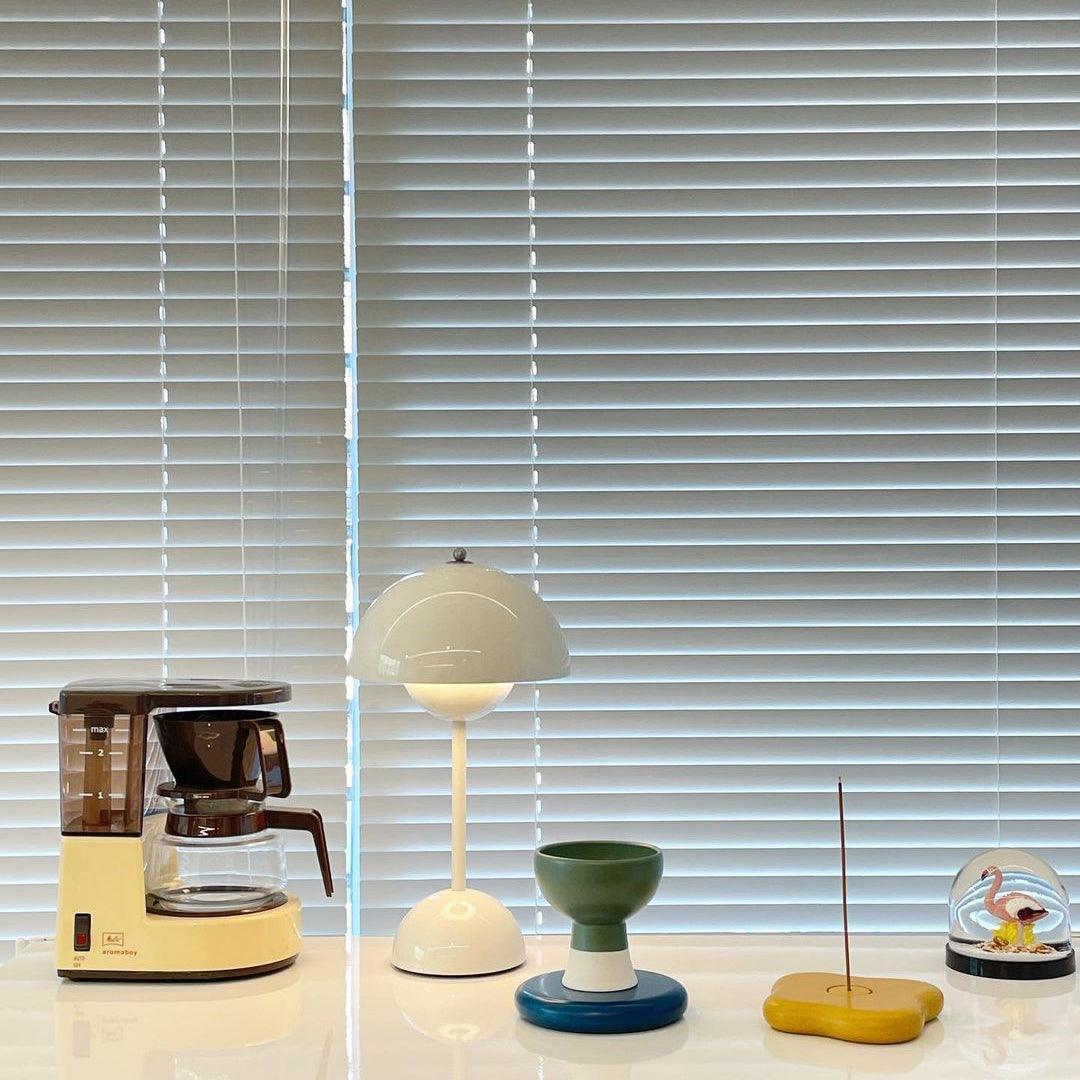 A pale cream Clérachic table lamp lit on a white shelf, beside a coffee maker and small coloured ornaments.