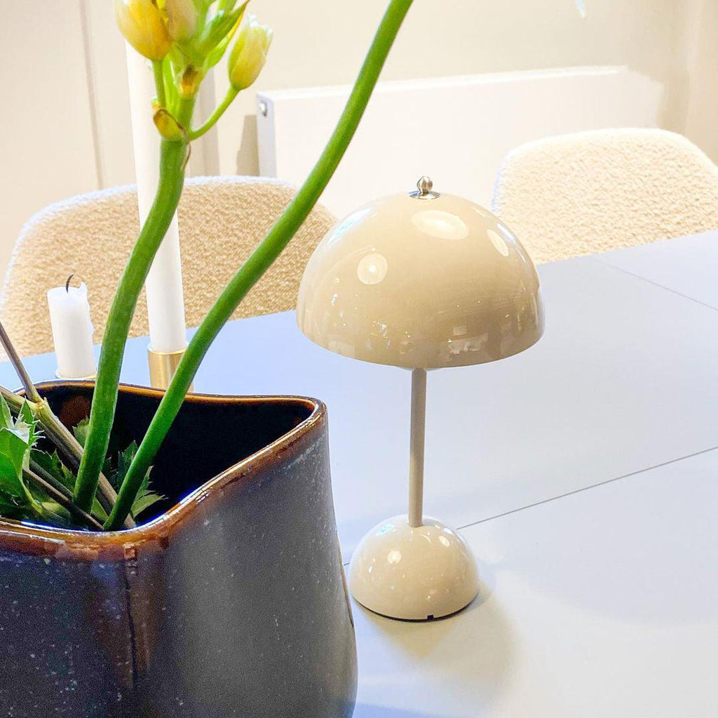 A cream Clérachic table lamp on a white table, with tall yellow flowers in a blue vase leaning in from the left.