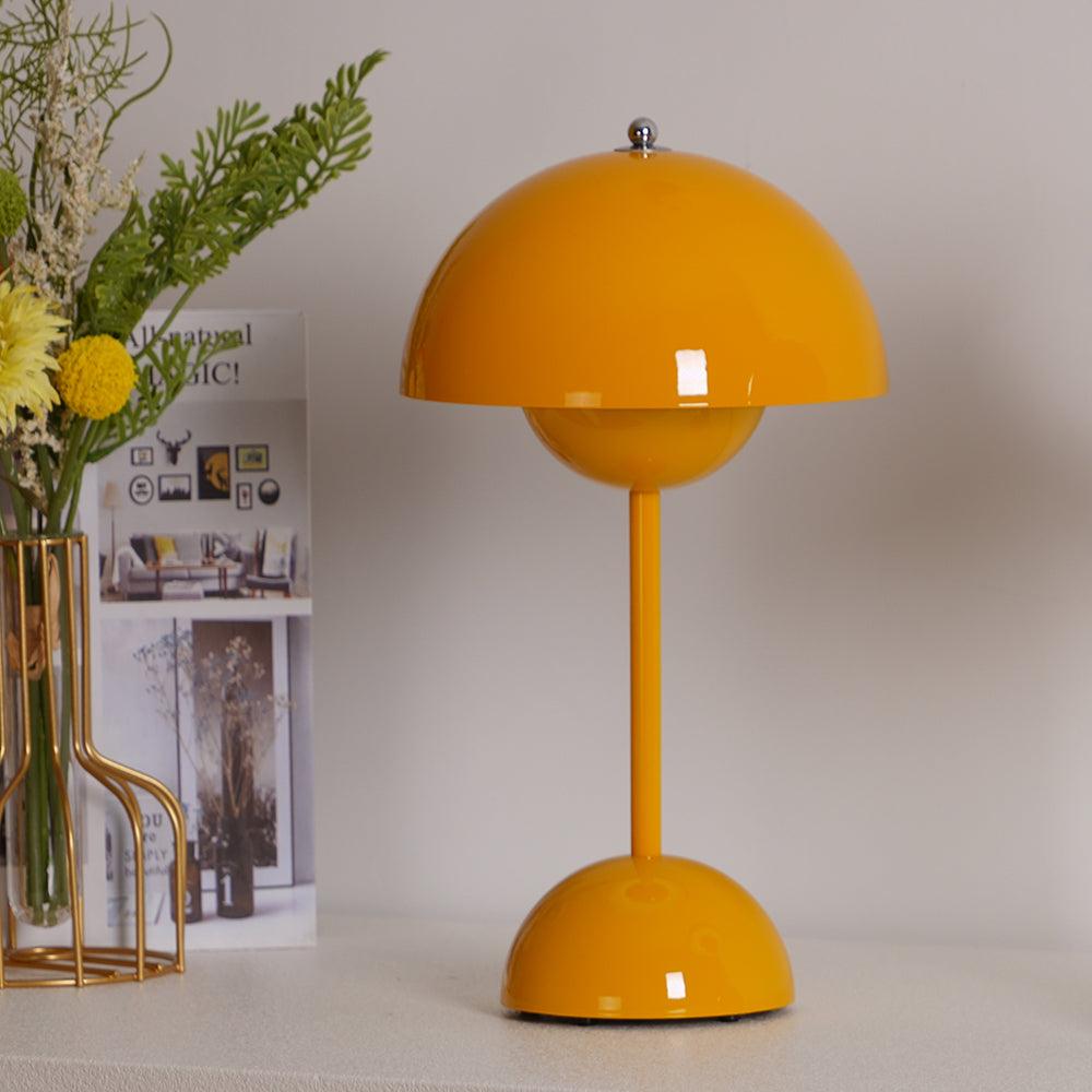 A yellow Clérachic table lamp on a pale surface, with dried flowers and a brass vase stand to the left.