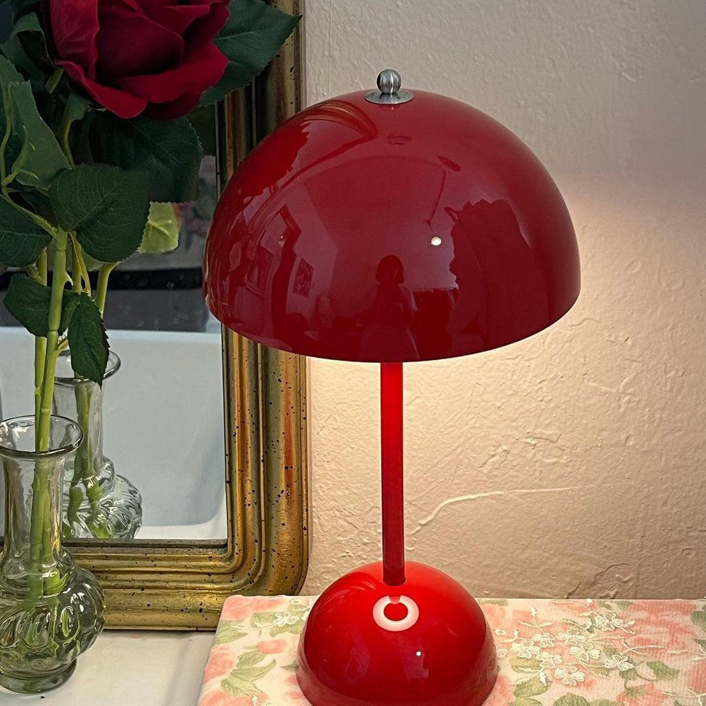 This Clérachic table lamp in glossy red, lit, in front of a gilt-framed mirror and a floral cloth.