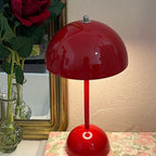 This Clérachic table lamp in glossy red, lit, in front of a gilt-framed mirror and a floral cloth.