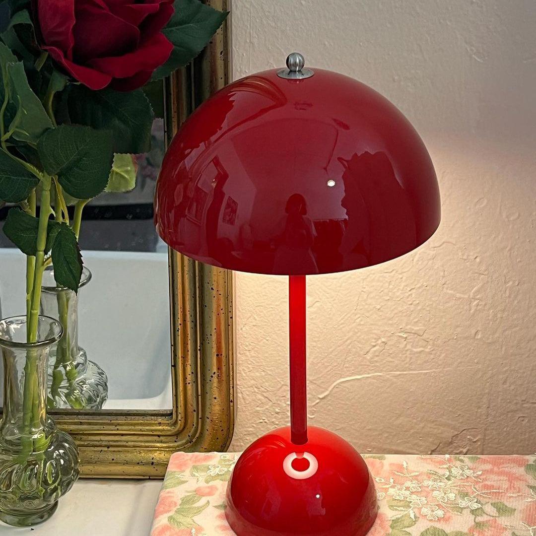This Clérachic table lamp in glossy red, lit, in front of a gilt-framed mirror and a floral cloth.