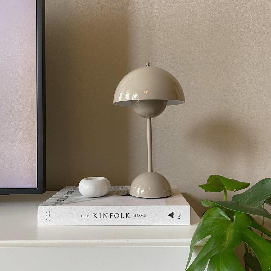 A greige Clérachic table lamp on a white shelf, beside a coffee-table book, a small white dish and a monstera leaf.