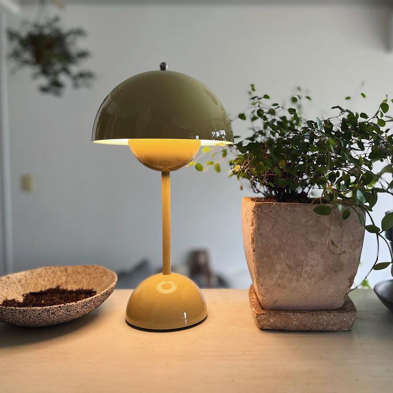 A lit olive-green Clérachic table lamp on a shelf, beside a potted plant and a shallow wooden bowl.