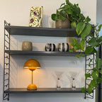 A yellow Clérachic table lamp lit on a dark wall shelf, with cocktail glasses and a plant, on a white mount.