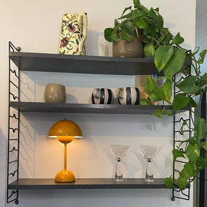 A yellow Clérachic table lamp lit on a dark wall shelf, with cocktail glasses and a plant, on a white mount.