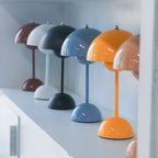 A row of Clérachic table lamps in plum, white, black, blue and orange, standing on a white shelf.
