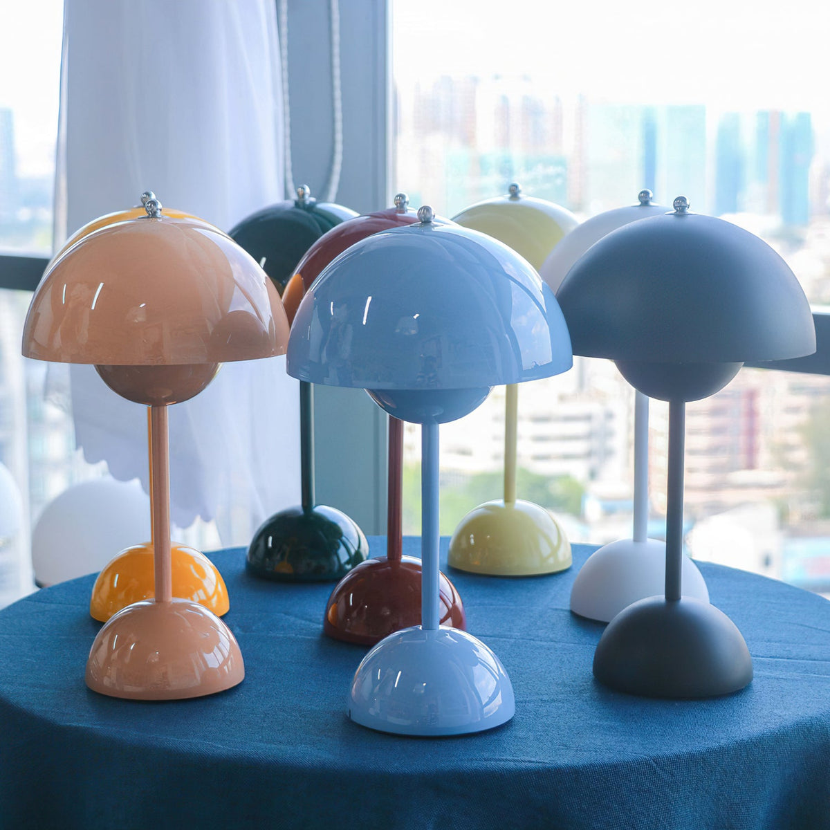 A group of Clérachic table lamps in blue, peach, green and grey on a blue cloth in front of a city window.