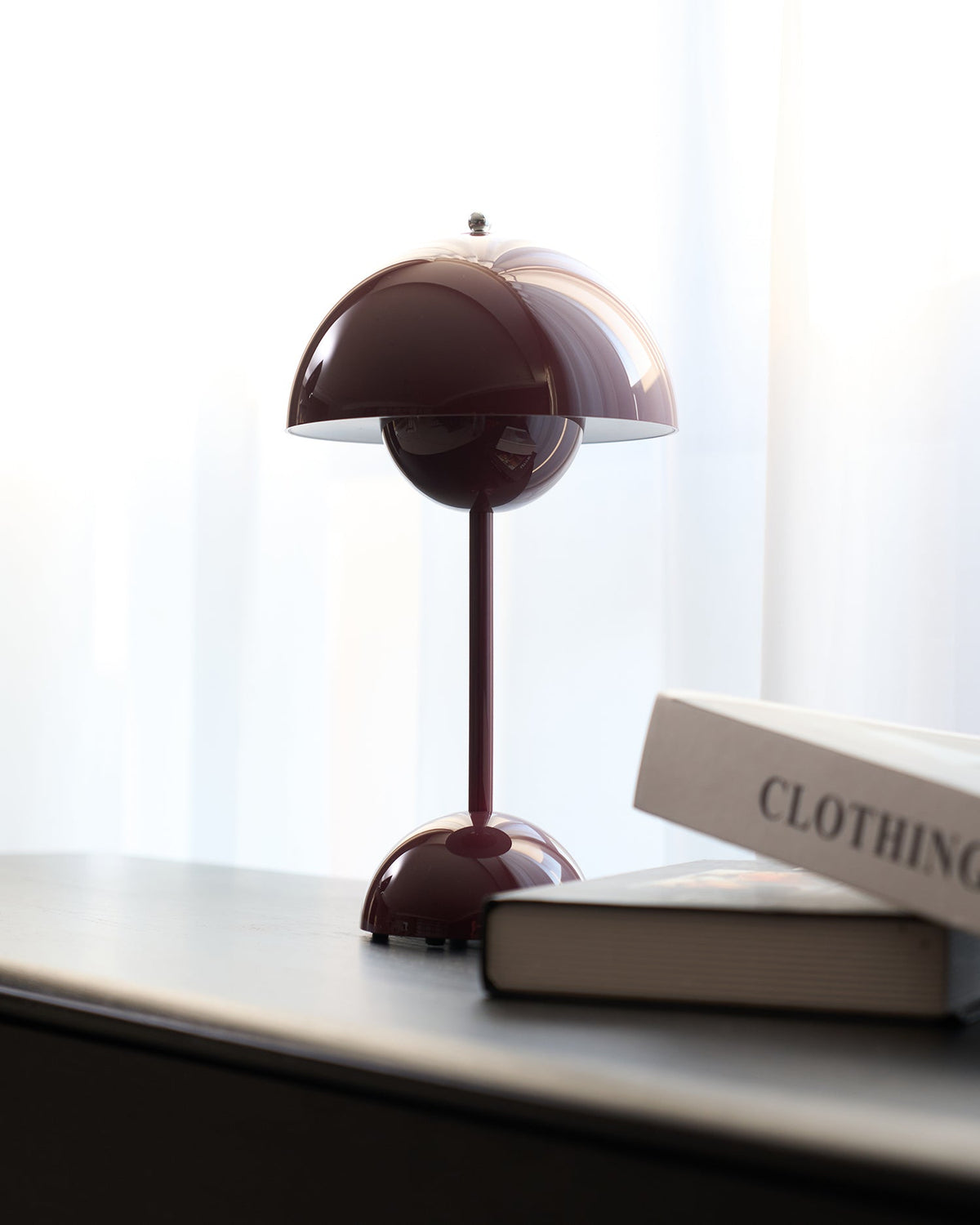 A deep plum Clérachic table lamp on a black desk beside two books, with a pale curtain behind it.