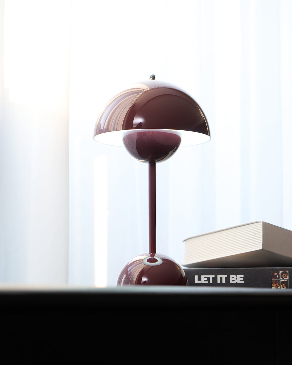 A deep plum Clérachic table lamp lit, with a bright rim under the shade, on a black desk beside books.